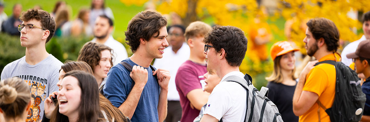 Two students talking outside in a crowd with backpacks. One is wearing a blue t-shirt and the other a white t-shirt.