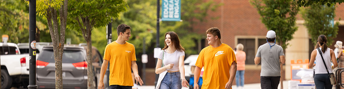 Three students walking. Two on the outside are wearing orange T-shirts while the middle is wearing a grey top with blue jeans.